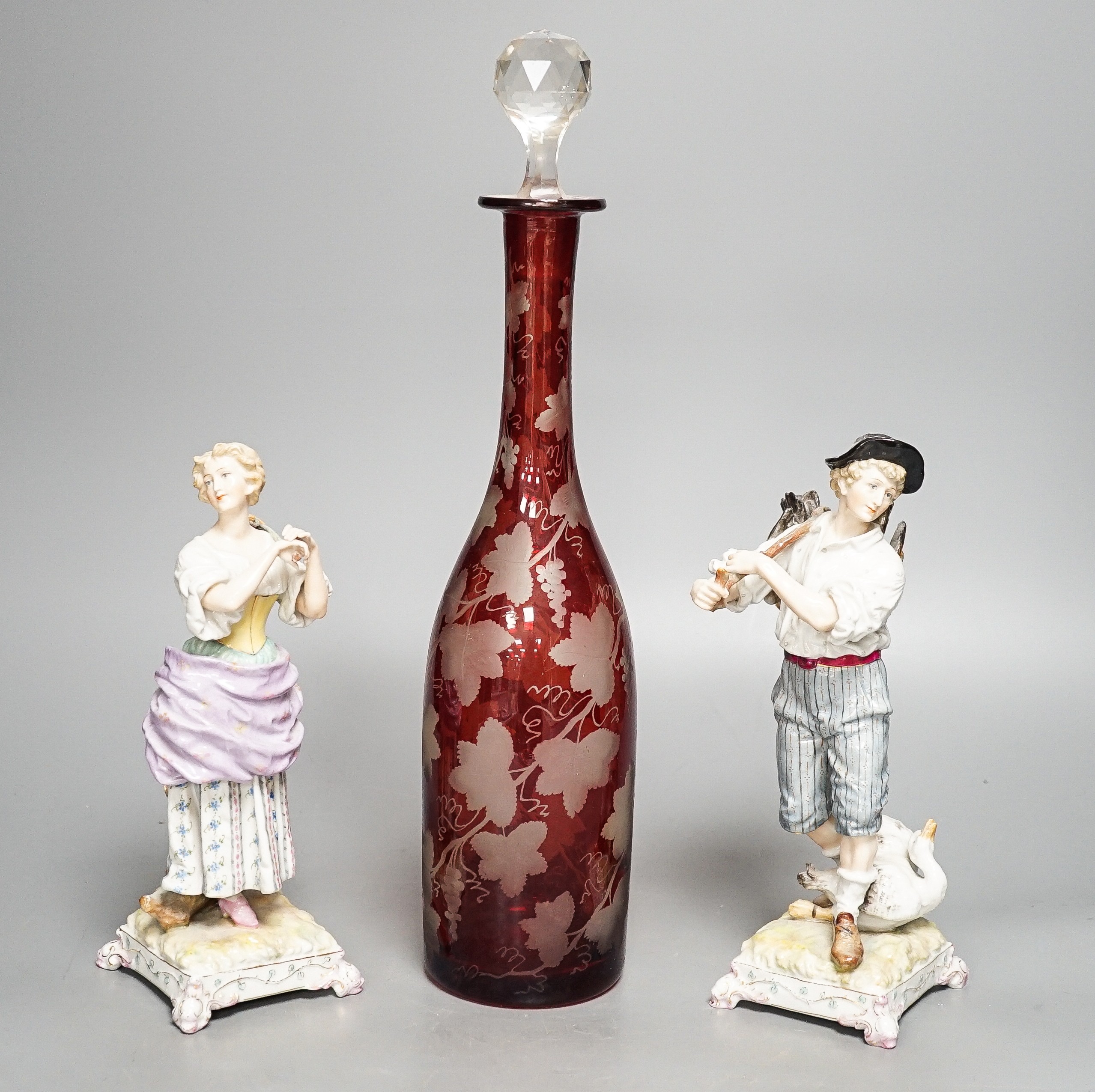 A late 19th century Bohemian wheel engraved ruby glass decanter and a pair of continental porcelain figures, decanter 37 cms high, including stopper.